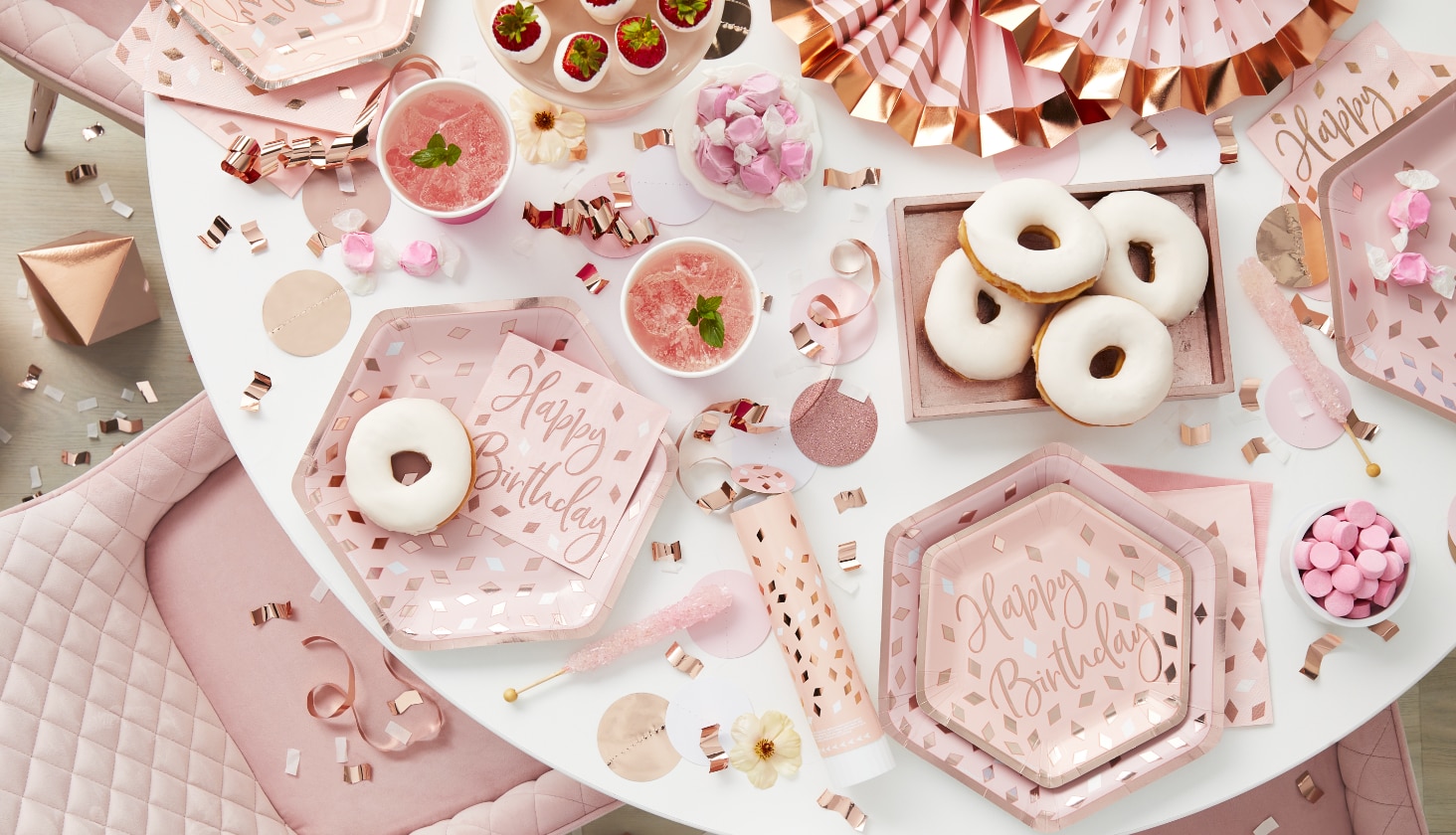 Une table décorée d’assiettes roses pour une fête d’anniversaire de naissance, de serviettes de table, de confettis, de boissons et de beignes blancs.