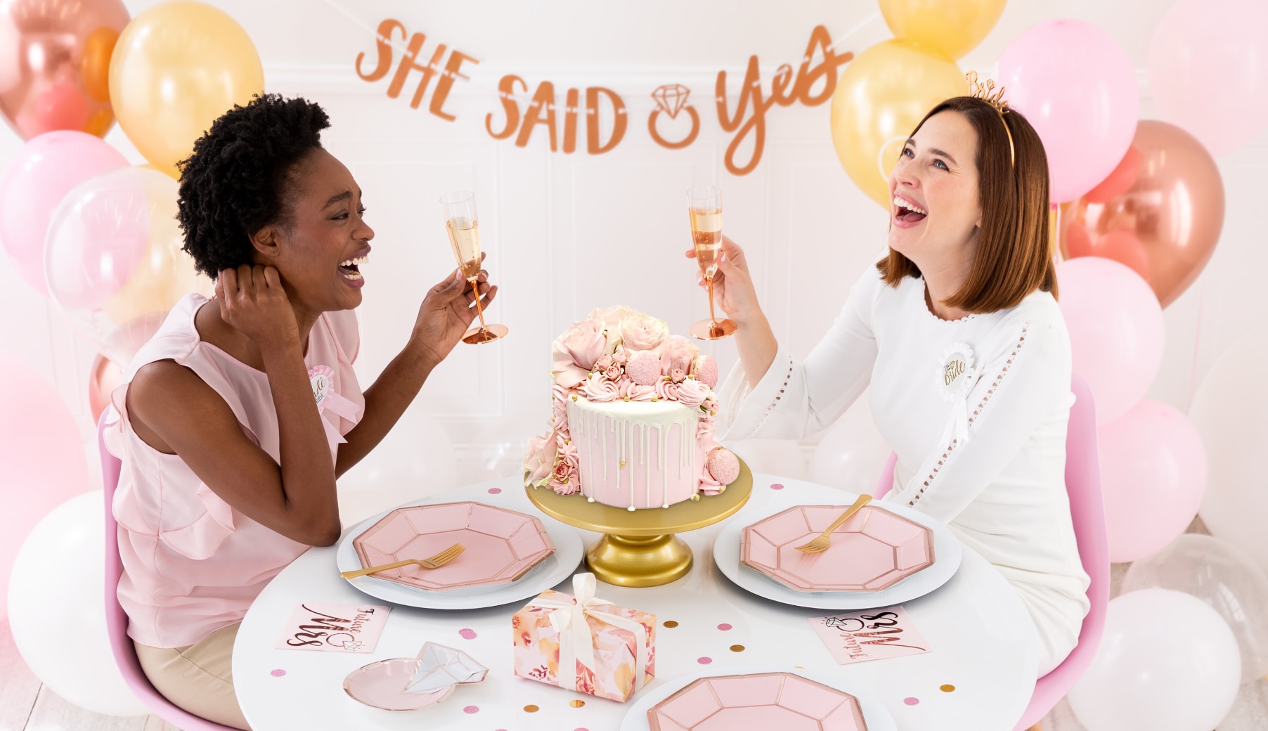 Two laughing women holding champagne glasses sitting at a table, in a room decorated with pink balloons, tableware and a banner that reads "SHE SAID YES".