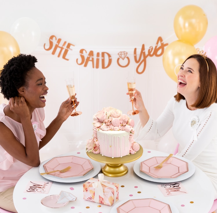 Two laughing women holding champagne glasses sitting at a table, in a room decorated with pink balloons, tableware and a banner that reads "SHE SAID YES".