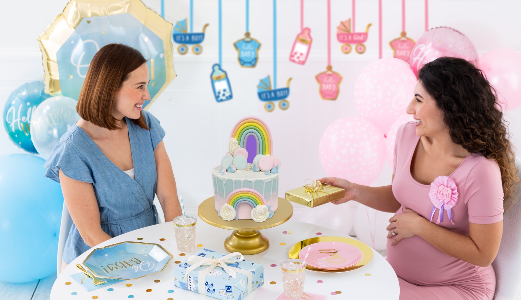 A pregnant woman and a friend sitting at a table in a room decorated in a pink and blue baby shower theme.