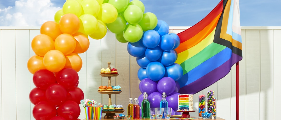A rainbow balloon arch, a Pride flag and a table decorated with assorted  rainbow-themed candy and cakes.