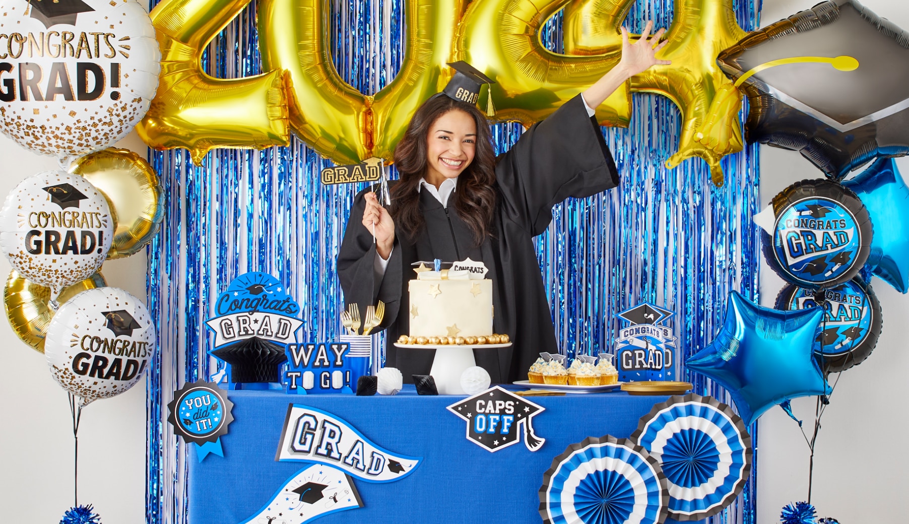 A woman wearing a graduation cap and gown while holding a sign that says 'grad', surrounded by graduation themed balloons and table decor.