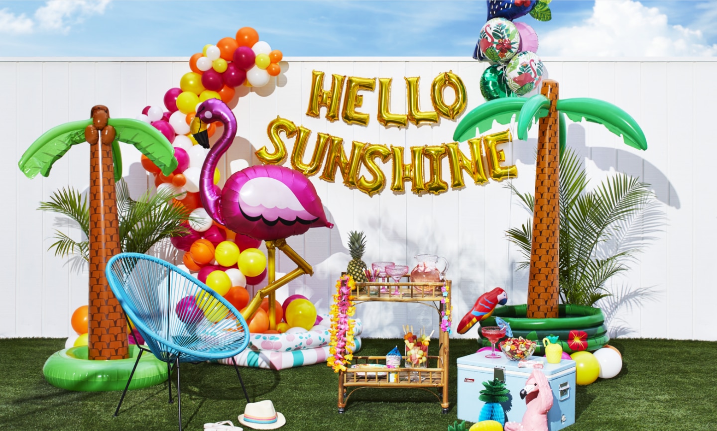 An outdoor space decorated with palm tree inflatables, a flamingo balloon, a balloon bouquet and a balloon banner that reads 'HELLO SUNSHINE'.