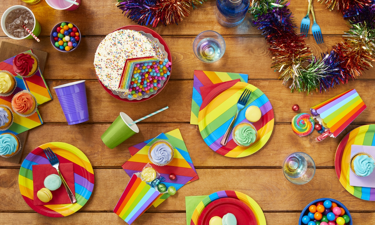 An overhead shot of various rainbow-themed tableware, cupcakes, cookies, candy and a cake.