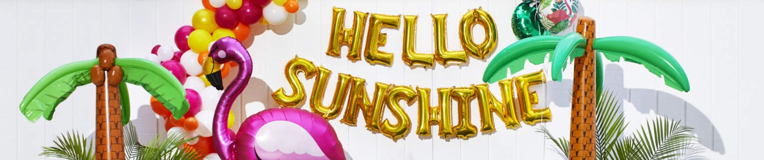 An outdoor space decorated with palm tree inflatables, a flamingo balloon, a balloon bouquet and a balloon banner that reads 'HELLO SUNSHINE'.
