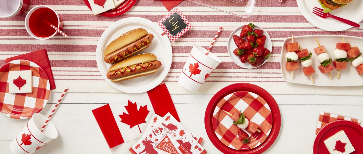An overhead shot of various red and white Canada Day themed tableware, hotdogs, strawberries and kebabs.