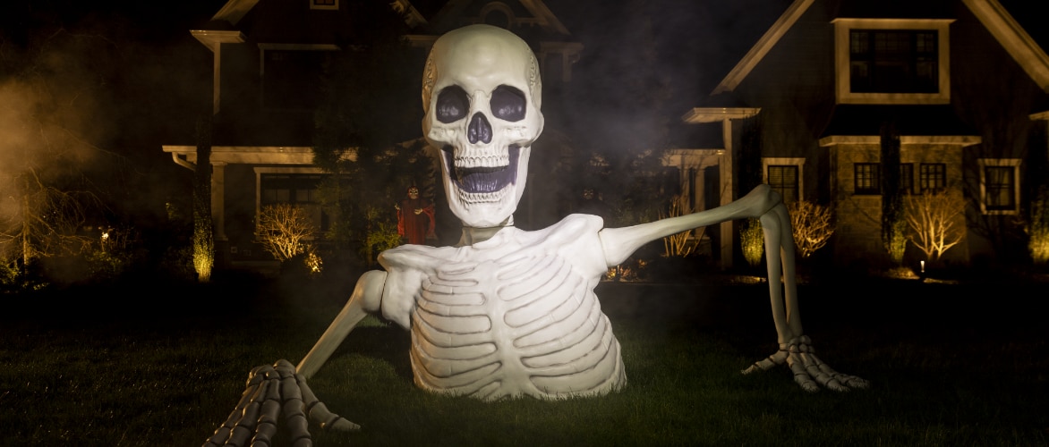 A giant skeleton  breaking out of the ground of a lawn decorated for Halloween.