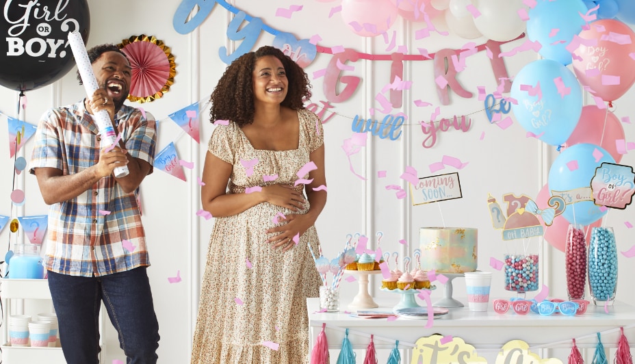 Un homme et une femme faisant éclater une bombe à confettis dans une pièce remplie d’articles de fête prénatale roses et bleus.