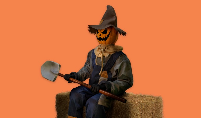 A pumpkin scarecrow sitting on a bale of hay holding a shovel.