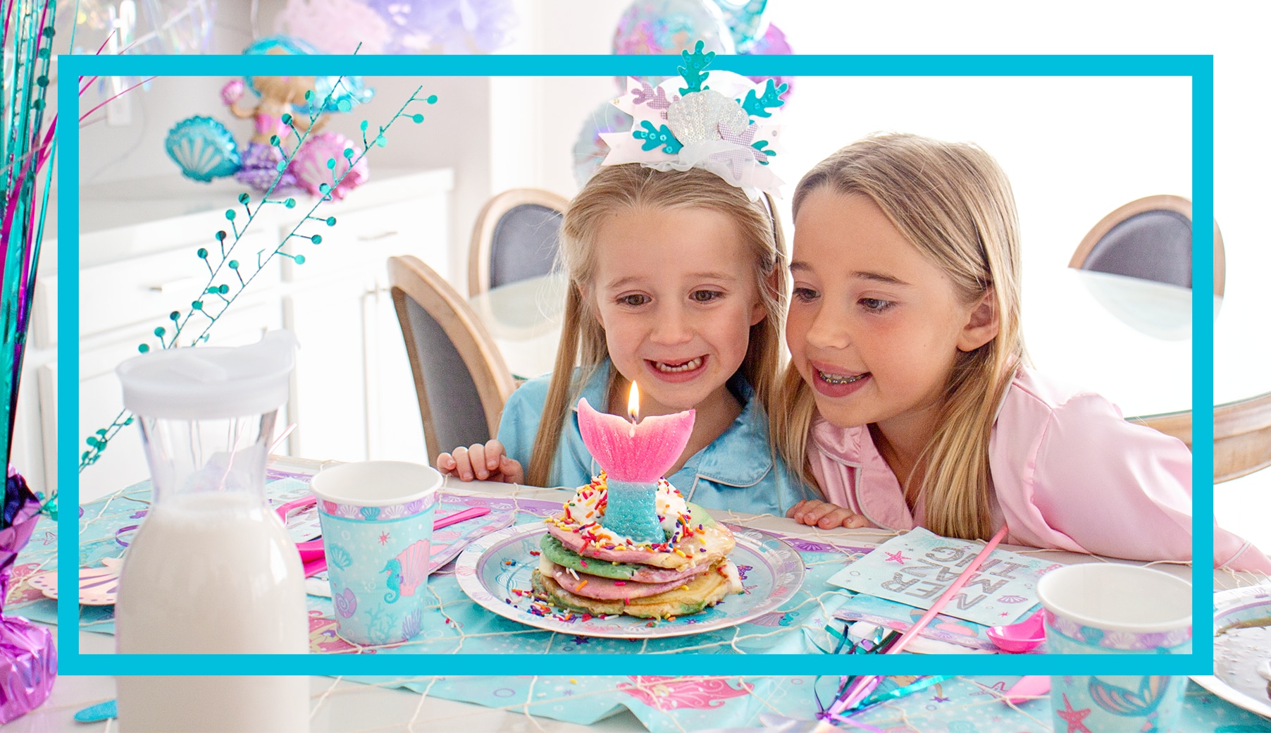 Two girls sitting at a table in front of a stack of pancakes with a mermaid tail candle on a table decorated for a birthday party. 