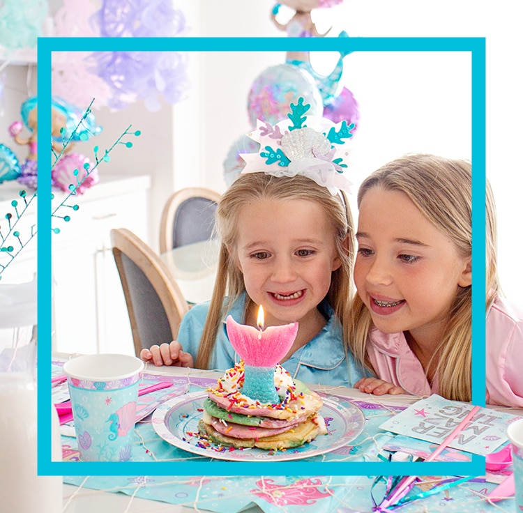 Two girls sitting at a table in front of a stack of pancakes with a mermaid tail candle on a table decorated for a birthday party. 