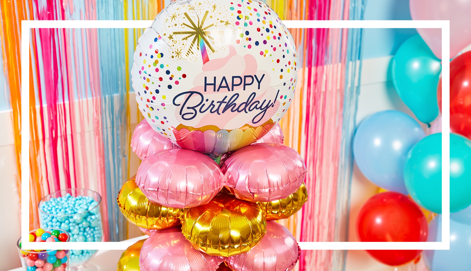 A white and multi-coloured helium-filled balloon with the words 'Happy Birthday' printed on the front stacked on top of pink and gold balloons.