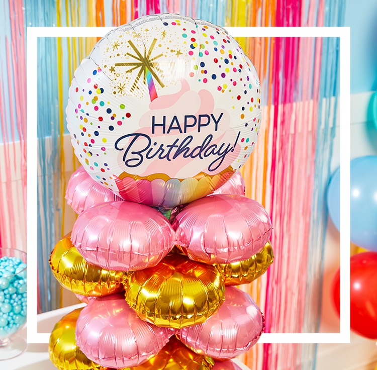 A white and multi-coloured helium-filled balloon with the words 'Happy Birthday' printed on the front stacked on top of pink and gold balloons.