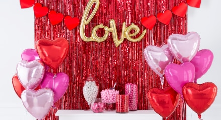 A red fringe, tinsel hanging wall decoration paired with a heart banner and a helium-filled balloon that spells the word 'Love' in a room decorated with pink and red balloons for Valentine's Day.