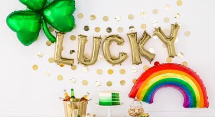 A bucket filled with champagne bottles sitting on a table beside a green cake with a gold helium-filled balloon on the wall behind it that says 'Lucky' beside a rainbow and four leaf clover balloon.