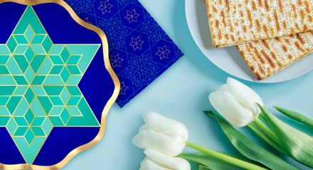 A navy blue, gold and green plate beside a bouquet of white tulips and a plate with food for Passover.