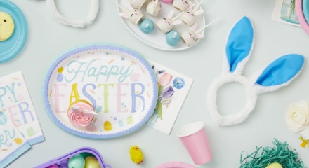 A pastel-coloured plate with the words 'Happy Easter' printed on it and a cupcake sitting on top, beside a pair of bunny ears, a pink solo cup and various Easter-themed crafts.