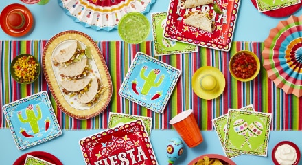 A bright and multi-colour table spread with tacos, salsa, guacamole, sombrero decorations, red plates with the word 'Fiesta' written on the front and other Mexican-themed tableware and decor.