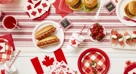 A red and white table spread decorated for Canadian Day with maple leaf-themed treats and cups, beside mini Canadian flags and plates with hotdogs, strawberries and burgers.