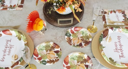 A table decorated in earth tones with white and gold plates that say Happy Thanksgiving' beside autumn leaves-themed side plates, an orange and gold turkey decoration and Thanksgiving centrepiece.