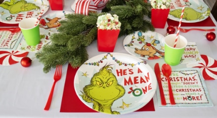 A table decorated for a Grinch-themed Christmas with red, mini popcorn holders, green paper cups, candy cane style straws, Grinch dinner plates and red cutlery.