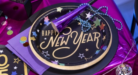 A black and gold plate with multi-coloured stars and gold font with the word 'Happy New Year' on the front beside purple napkins and horn, blue beads and mini disco balls.