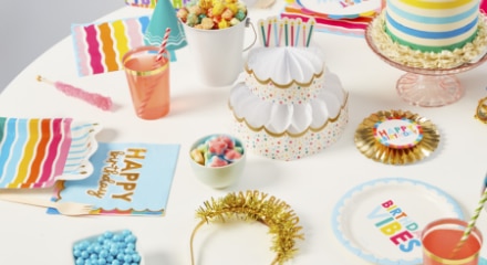 A white table decorated with gold accents and multi-coloured birthday decorations, with napkins and plates that say 'Birthday Vibes' and 'Happy Birthday' printed on the front.