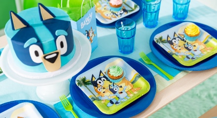 A table decorated for Bluey-themed kids' birthday party with a Bluey cake, dinner plates, favour bag and blue cups.