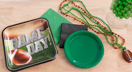 A football-themed party plate with the word 'Game Day' printed on the front beside black and green napkins, a green plate and colourful beaded necklaces with football charms.