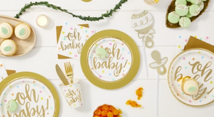 A white table decorated with sweet treats, white and gold napkins, plates and cups that say 'Oh Baby!' printed on the front for a baby shower.