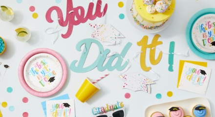 A white table with pink, yellow and blue letters that say 'You Did It!' with multi-coloured tableware, sweets and plates that say 'The Best Is Yet To Come' for a graduation party.