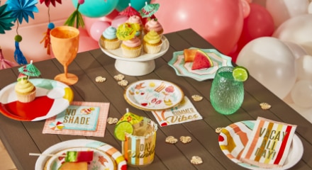 A wooden table surrounded by bright balloons, multicoloured summer-themed tableware and plates and napkins that say "Summer Vibes', 'Vacay All Day' and 'No Shade'.