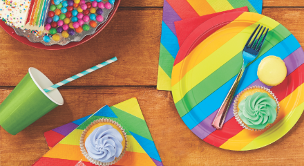 A wooden table with Pride-themed cake, napkins, plates and cupcakes.