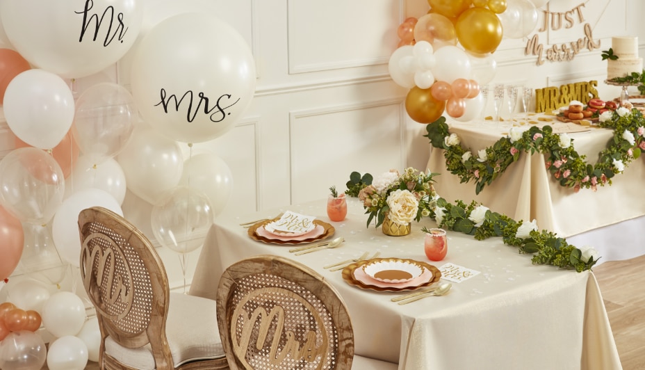 Deux tables dans une pièce blanche décorées pour un mariage avec des ballons blancs, dorés et roses sur lesquels sont écrits « Mr. and Mrs. ».