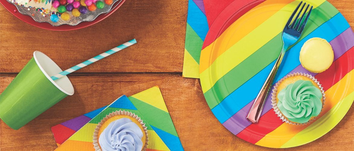 A colorful party setup with a green cup, striped straw, rainbow-striped plates and napkins, and assorted treats on a wooden table.