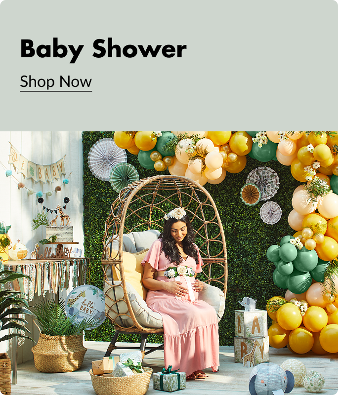 A pregnant woman in a pink dress and flower crown sitting in a wicker egg chair surrounded by green, yellow and peach balloons and baby shower decorations. 