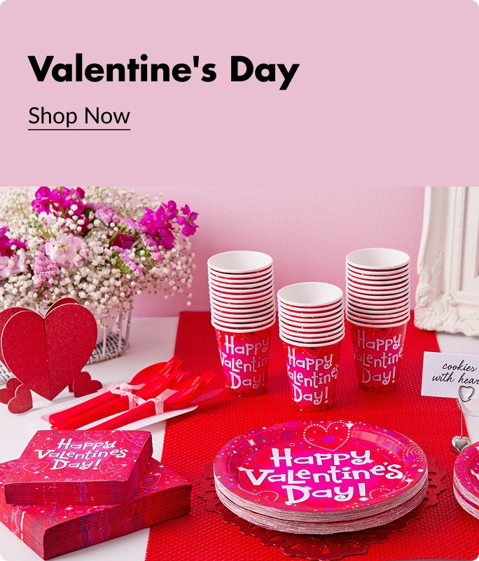 Pink and red plates, cups and napkins that say 'Happy Valentine's Day' printed on the front sitting beside a vase filled with flowers, heart decoration and cutlery on a red and white table. 